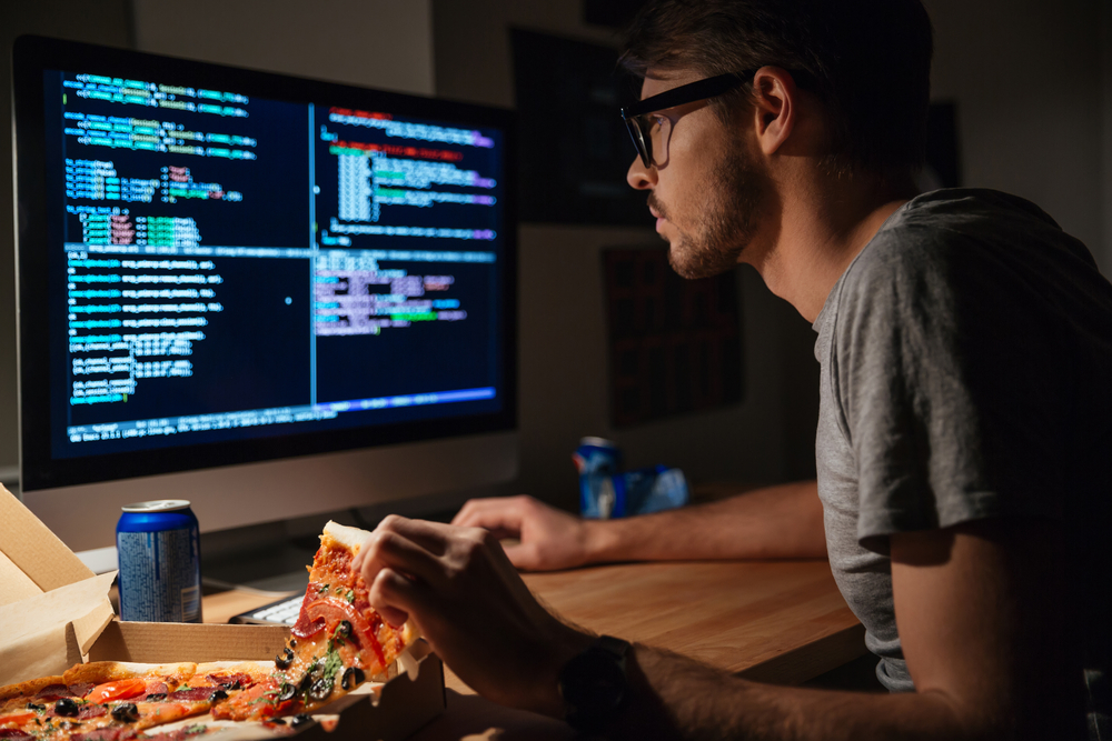 Profile of concentrated young software developer eating pizza and coding at home
